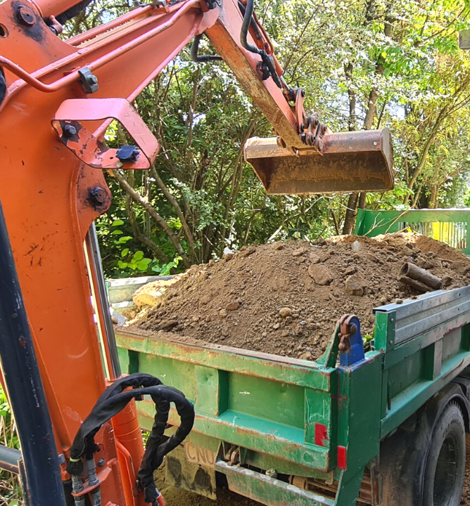 Orange Digger loading dirt into the back of a green truck on building site.